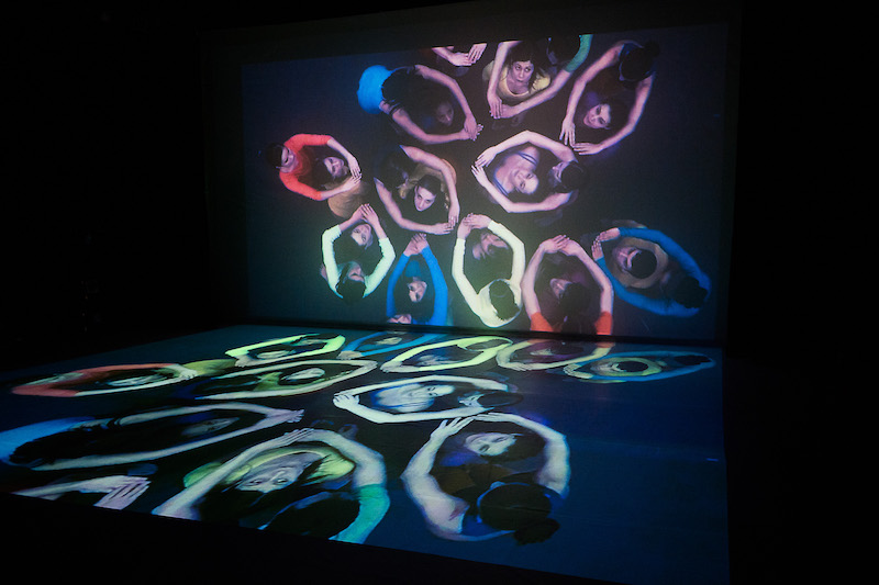 A film of a group of people in bright prints is projected on the back wall of the theater and on its floor